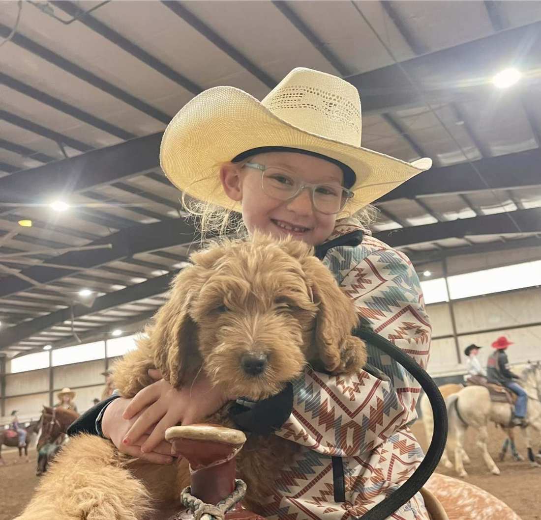One of the girls in a cowboy hat, on horseback in the arena, holding a Goldendoodle puppy